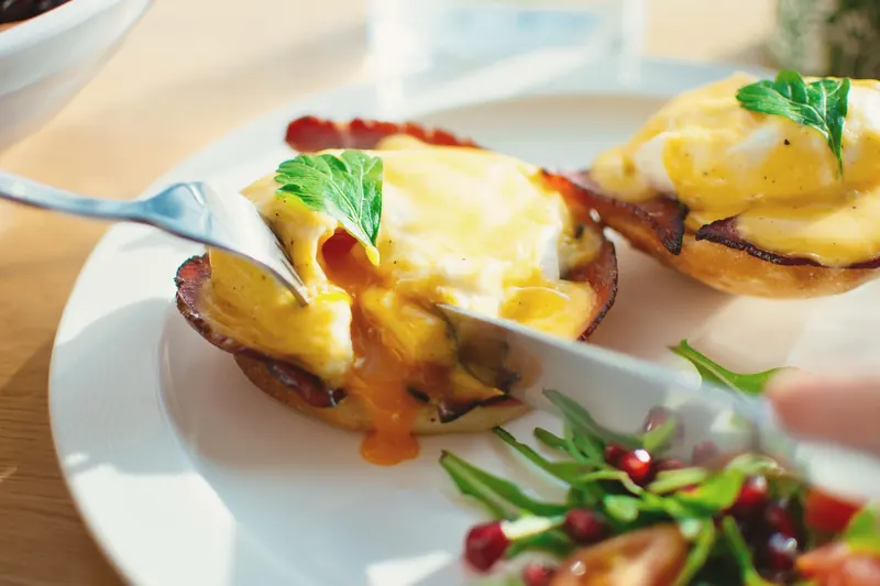 Fork and knife cutting into eggs Benedict with hollandaise, a rocket and pomegranate salad beside