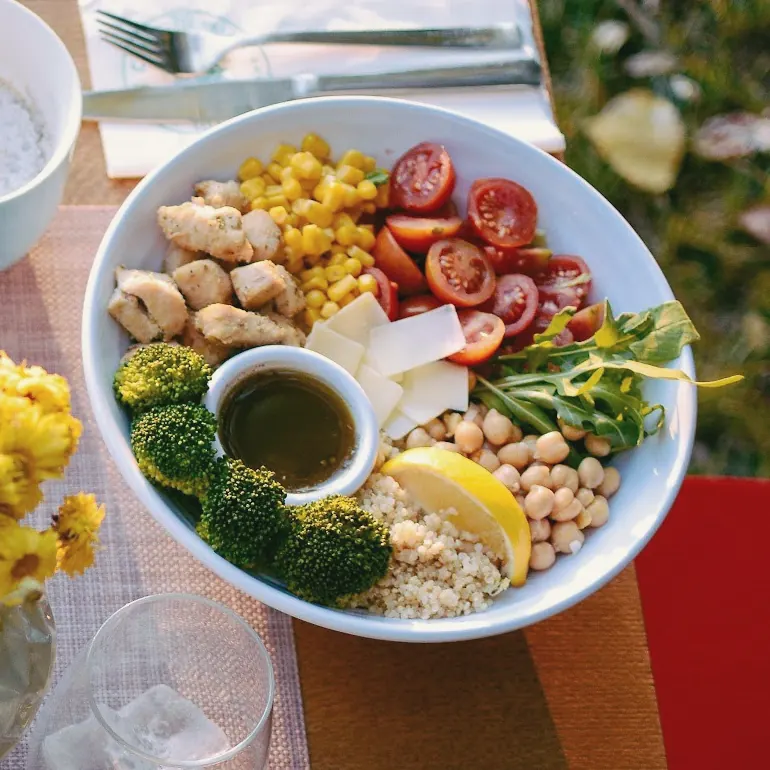 A bowl of chicken, corn, chickpeas, broccoli, quinoa and tomatoes with a dressing pot