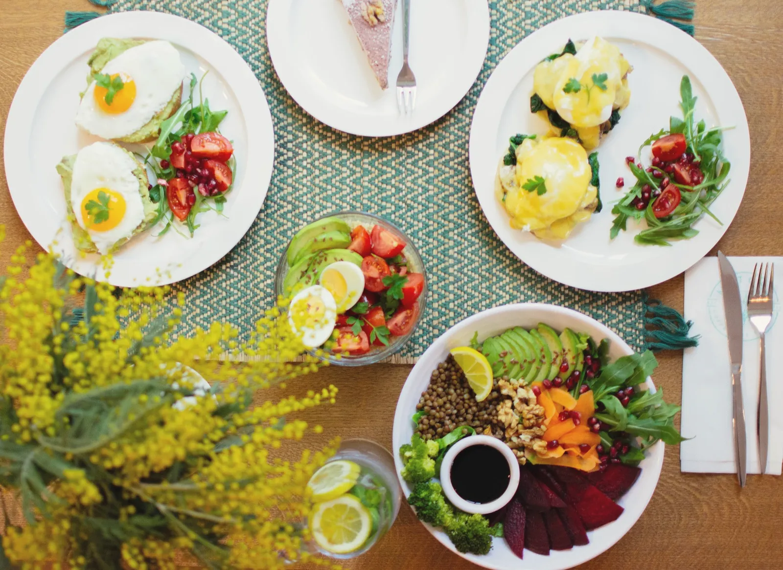 Brunch spread from above: eggs Benedict, fried eggs, a grain bowl and mimosa flowers